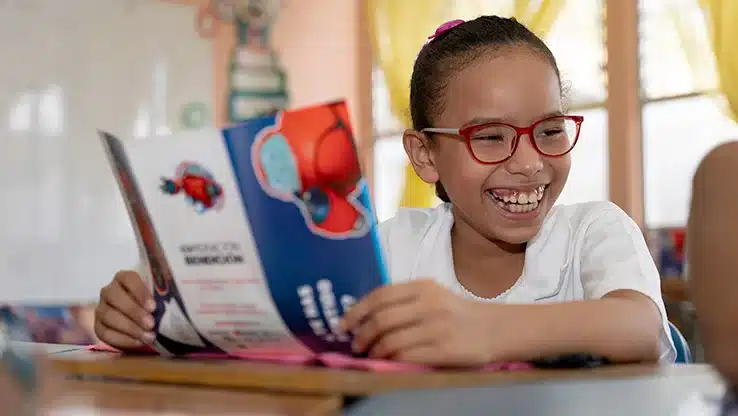 Smiling girl in red glasses holding a colourful Superbook booklet in a classroom setting