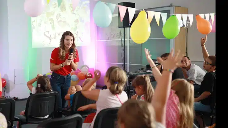 Female presenter in a red shirt engaging with children in a decorated room with balloons and a Superbook projection