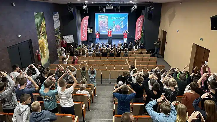 Children in an auditorium enthusiastically participating in a Superbook show, with presenters on stage and a vibrant screen display